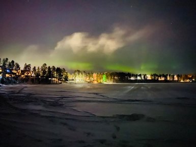 Ein winterlicher See unter einem sternenklaren Himmel, beleuchtet von den tanzenden Lichtern des Nordlichts.