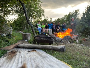 Eine Gruppe von Kindern sitzt um ein Lagerfeuer, während ein Erwachsener etwas in der Hand hält und ihnen zuschaut.