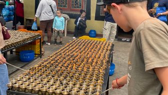 Ein Junge zündet Kerzen auf einem Tisch an, umgeben von zahlreichen brennenden Öllampen in einem geschäftigen Tempelbereich.