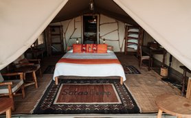 Ein gemütliches Zeltinnere mit einem großen Bett, bunten Kissen und einem Teppich mit der Aufschrift 'Satao Camp'.