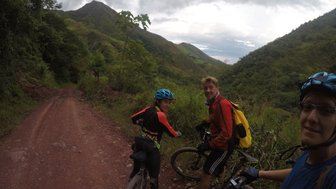 Drei Mountainbiker stehen auf einem schmalen, roten Pfad, umgeben von üppigem Grün und sanften Hügeln im Hintergrund.