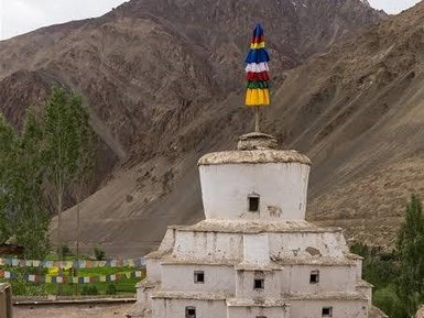 Ein weißer Stupa mit buntem Fahnenmast steht in einer bergigen Landschaft, umgeben von grünen Bäumen und Gebetsfahnen.