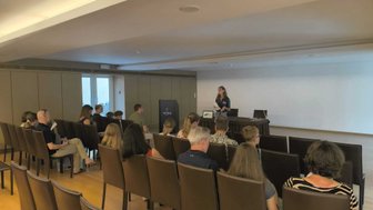 Eine Gruppe von Menschen sitzt in einem modernen Raum und hört einer Präsentation einer Frau am Pult zu.
