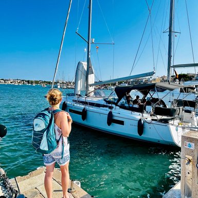 Eine Frau mit einem Rucksack steht am Hafen und beobachtet ein Boot, das im klaren Wasser anlegt.