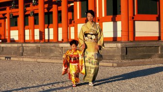 Eine Frau in einem eleganten Kimono geht mit einem kleinen Mädchen, das ebenfalls traditionell gekleidet ist, durch einen Tempelbereich.