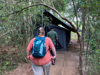 Eine Frau mit einem Rucksack geht auf einem schmalen Pfad zu einem Zelt, umgeben von üppigem Grün und Bäumen.