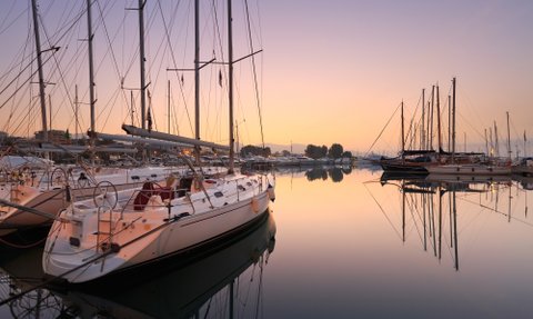 Eine ruhige Marina bei Sonnenaufgang, mit mehreren Segelbooten, die sanft im Wasser schaukeln und ihre Spiegelungen zeigen.