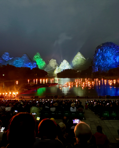 Eine beeindruckende Lichtshow erhellt die Karstberge, während Zuschauer am Ufer gebannt zuschauen und ihre Handys benutzen.
