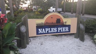 Ein farbenfrohes Schild mit der Aufschrift 'NAPLES PIER' steht vor einer üppigen, grünen Landschaft und Palmen.