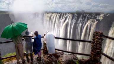 Eine Gruppe von Menschen steht am Geländer und bewundert die majestätischen Victoriafälle, während der Regenbogen im Nebel schimmert.