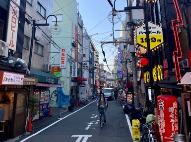 Eine belebte Straße in Osaka, gesäumt von bunten Geschäften und Restaurants, mit einem Radfahrer, der vorbeifährt.