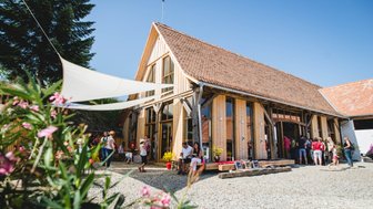 Eine lebhafte Szene vor einem modernen Holzgebäude, umgeben von Menschen, die sich unter einem blauen Himmel versammeln.