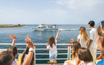 Eine Gruppe von Passagieren winkt einem Boot zu, das sanft über das Wasser gleitet, während die Sonne am Himmel scheint.