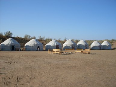 Sechs traditionelle Jurten stehen in einer trockenen, sandigen Landschaft unter klarem, blauem Himmel.