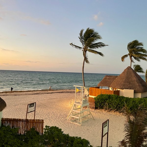 Ein ruhiger Strand mit sanften Wellen, umgeben von Palmen und traditionellen Strohdächern, während die Sonne untergeht.