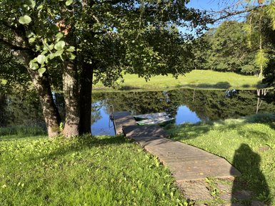 Ein ruhiger See spiegelt die umgebende Natur wider, während ein Holzsteg zu einem Boot führt, das am Ufer liegt.