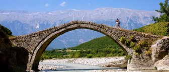 Ein Paar geht über eine alte Steinbrücke, umgeben von grünen Hügeln und majestätischen Bergen unter einem strahlend blauen Himmel.