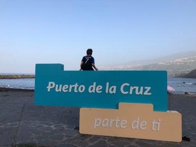 Ein Junge sitzt auf einem bunten Schild mit der Aufschrift 'Puerto de la Cruz' am Strand, während das Meer im Hintergrund glitzert.