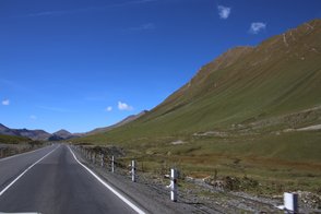 Eine kurvenreiche Straße schlängelt sich durch eine grüne Landschaft, umgeben von majestätischen Bergen unter einem klaren blauen Himmel.