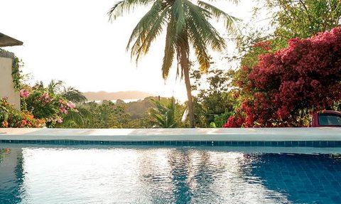 Ein glitzernder Pool spiegelt die Farben des Sonnenuntergangs wider, umgeben von üppigem Grün und blühenden Pflanzen.