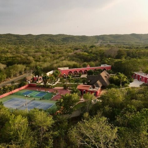 Eine Luftaufnahme einer farbenfrohen Hacienda mit Tennisplätzen, umgeben von üppiger, grüner Vegetation und sanften Hügeln.