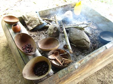 Ein rustikales Kochfeld mit dampfenden Steinen und Holz, umgeben von Schalen mit Kakaobohnen und Werkzeugen zum Zubereiten von Schokolade.