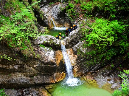 Eine Gruppe von Abenteurern steht an einem kleinen Wasserfall in einer grünen Schlucht, umgeben von üppiger Vegetation.