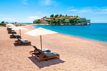 Ein sonniger Strand mit Liegen und Sonnenschirmen, die auf das klare, blaue Wasser blicken, umgeben von einer malerischen Küstenlandschaft.