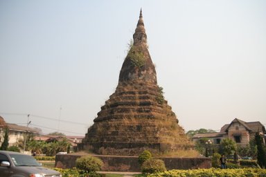 Eine alte, mit Gras bewachsene Stupa erhebt sich majestätisch in der Stadt, umgeben von modernen Gebäuden und Bäumen.