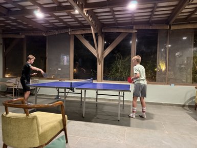 Zwei Jungen spielen Tischtennis in einem gemütlichen Raum mit großen Fenstern und sanfter Beleuchtung.