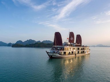 Ein elegantes Kreuzfahrtschiff mit braunen Segeln gleitet sanft über das ruhige Wasser der Halong-Bucht, umgeben von grünen Inseln.