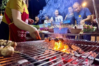 Eine Person grillt Fleischspieße über einem offenen Feuer, während Rauch in die Luft steigt und hungrige Gäste zuschauen.