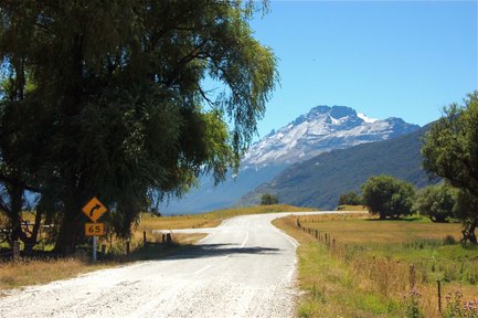Eine kurvenreiche Straße führt durch eine malerische Landschaft mit schneebedeckten Bergen im Hintergrund und üppigem Grün.