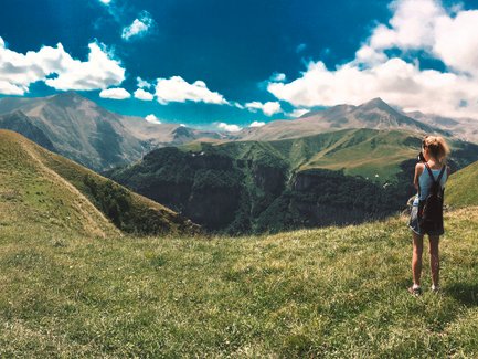 Eine Person steht auf einer grünen Wiese und blickt auf majestätische Berge unter einem strahlend blauen Himmel.