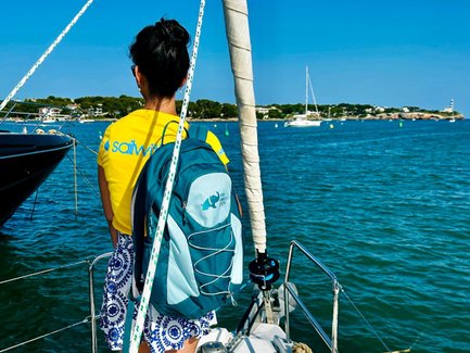 Eine Person mit einem Rucksack steht auf einem Segelboot und blickt auf das ruhige Wasser und die umliegende Landschaft.