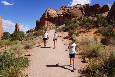 Eine Familie wandert auf einem schmalen Pfad im Arches Nationalpark, umgeben von beeindruckenden roten Felsen und grünem Gestrüpp.