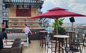 Eine stilvolle Dachterrasse mit einer Bar, umgeben von Pflanzen und einem Blick auf die Stadt, während ein Kellner vorbeigeht.