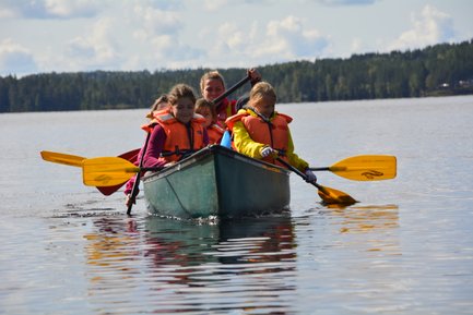 Vier Kinder in einem Kanu paddeln fröhlich auf einem ruhigen See, umgeben von grünen Bäumen und einem klaren Himmel.
