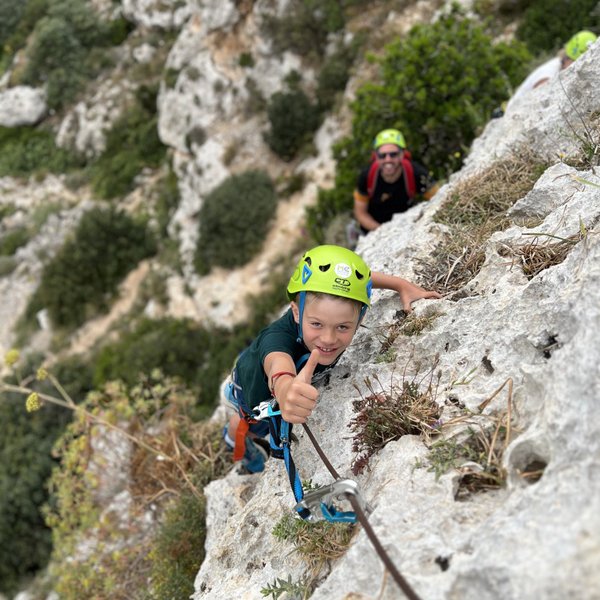 Ein Junge mit einem grünen Helm klettert an einer steilen Felswand und zeigt ein Daumen-hoch-Zeichen.