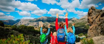 Eine Gruppe von vier Personen, darunter Kinder, genießt die Aussicht auf eine beeindruckende Berglandschaft und hebt die Hände.