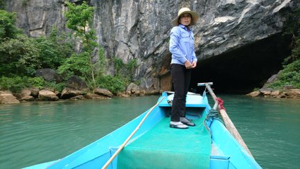 Eine Frau steht auf einem blauen Boot, umgeben von grünen Bäumen und steilen Felsen, während sie in eine Höhle blickt.