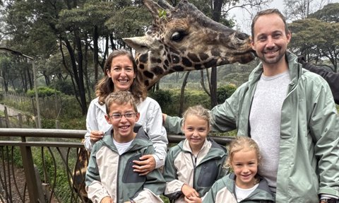 Eine Familie steht auf einer Holzterrasse, während eine Giraffe neugierig in die Kamera schaut und die Kinder lächeln.