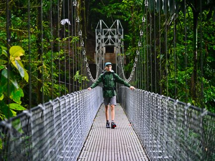 Ein Wanderer in einem lebhaften grünen Mantel steht gelassen auf einer Hängebrücke, umgeben von üppiger tropischer Vegetation.