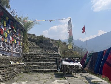 Eine steinerne Treppe führt zu einem Aussichtspunkt, umgeben von bunten Gebetsfahnen und einem klaren blauen Himmel.