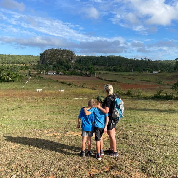 Eine Frau steht mit zwei Kindern auf einer Wiese und blickt auf eine malerische Landschaft mit sanften Hügeln.