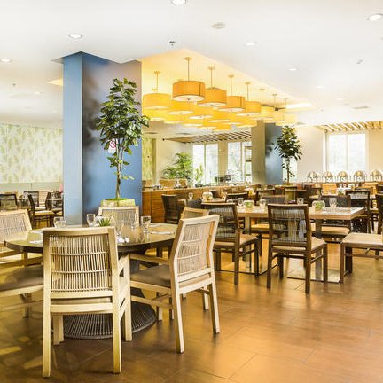 Ein helles Restaurant mit modernen Holztischen und Stühlen, umgeben von Pflanzen und großen Fenstern, die Tageslicht hereinlassen.