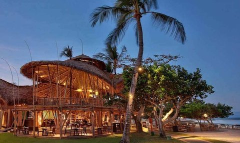 Ein stilvolles Restaurant mit Strohdach, umgeben von Palmen und sanftem Licht, das eine entspannte Atmosphäre schafft.