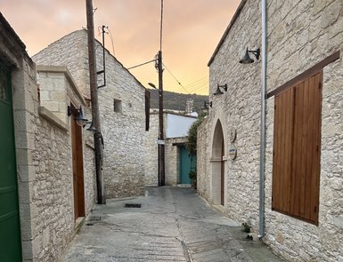 Eine ruhige Gasse mit steinernen Wänden und Holztüren, beleuchtet von sanften Straßenlaternen, unter einem orangefarbenen Himmel.