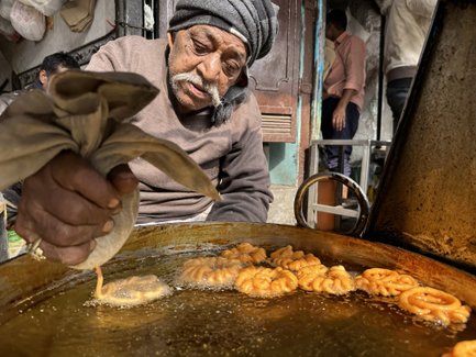 Ein älterer Mann bereitet frisch frittierte Süßigkeiten in einer großen Pfanne auf einem belebten Markt in Delhi zu.