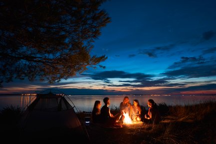 Eine Gruppe von fünf Freunden sitzt um ein Lagerfeuer am Ufer, während der Sonnenuntergang den Himmel in warmen Farben erleuchtet.